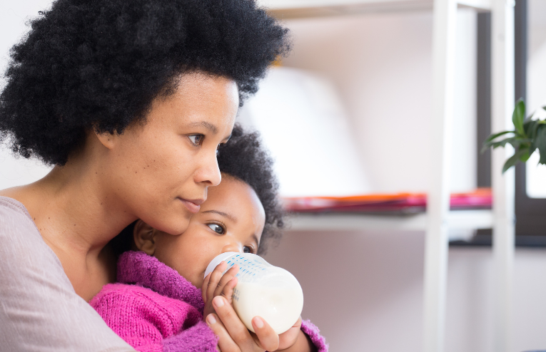Mother and child with brown skin and curly hair skin as mother holds child drinking a bottle.