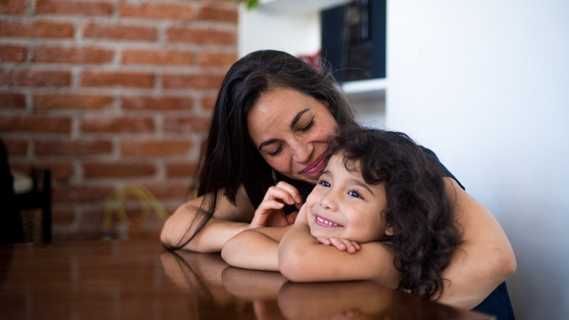 Woman and child sit at a table, both with light brown skin and dark curly hair. Woman's arm is around smiling child.